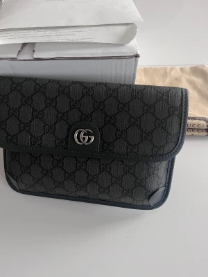 Svart axelväska från Gucci med GG-mönster - Snygg svart axelväska från Gucci med klassiskt GG-mönster över hela väskan. Väskan har en justerbar rem med spänne och silverfärgad GG-logga framtill. Materialet är en mix av skinn och syntet, perfekt för dig som gillar stilrena designer och exklusiva detaljer.