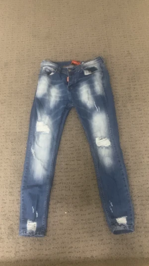 Blå Dsquared2 skinny jeans med slitningar - Säljer ett par blå skinny jeans från Dsquared2 med coola slitningar och blekta detaljer på både fram- och baksida. Jeansen har klassisk femficksdesign, låg midja och orange Dsquared2-märke bak. Perfekt för dig som gillar streetstyle och edgy look.