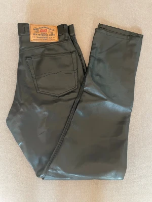 Svarta skinnbyxor från MGB Jeanswear - Svarta byxor i skinnimitation från MGB Jeanswear med klassisk femficksdesign och raka ben. Snyggt läderlook med traditionell jeanspatch bak i midjan. Perfekt för dig som vill sticka ut med en cool och edgy stil. Mått midj tvärs över 33,5 cm, innerben 75 cm