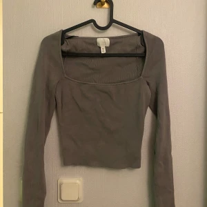 Grå ribbad långärmad topp från H&M - Säljer en grå croppad långärmad topp från H&M med fyrkantig halsringning. Toppen är ribbad och har en slim passform som sitter snyggt på kroppen. Perfekt att styla med jeans eller kjol för en clean look.