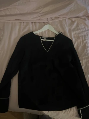Svart blus med vit kant från H&M - Snygg svart blus från H&M i storlek 36 med vit kant längs v-ringningen och ärmsluten. Blusen har lång ärm och en stilren, enkel design som passar perfekt till jeans eller kjol. Materialet känns mjukt och lätt mot huden.
