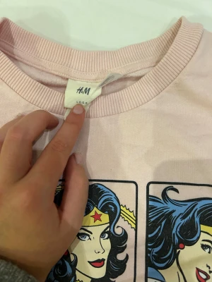Rosa Wonder Woman sweatshirt från H&M - Säljer en ljusrosa sweatshirt från H&M med coolt Wonder Woman-tryck i serietidningsstil på framsidan. Tröjan har ribbade muddar vid hals, ärmslut och nederkant samt lång ärm. Materialet är mjukt, perfekt för dig som gillar superhjältar och retro vibes.
