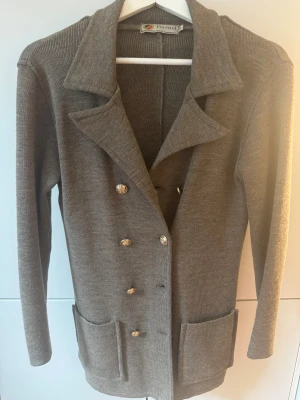 Brun/Grå cardigan jacka från Busnel - Snygg brun/grå cardigan jacka från Busnel med guldknappar och två stora fickor framtill. Jackan är stickad i ull och har klassisk krage samt lång ärm med Busnel-logga på mudden. Perfekt för dig som gillar stilrena och tidlösa plagg. Väldigt fint skick.
