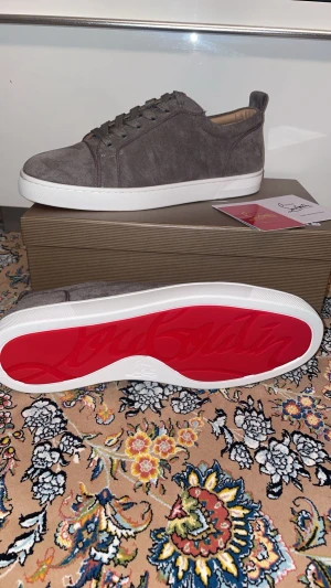Christian Louboutin grå mockasneakers - Säljer ett par grå sneakers från Christian Louboutin i mjuk mocka med klassisk röd sula. Skorna har rund tå, snörning och vit platt sula. Perfekt för dig som vill ha en clean och lyxig look att ens försöka!