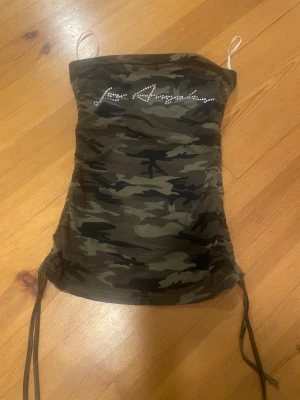 Camouflage tubtopp med snörning - Säljer en tubtopp med camouflage-mönster och snörning på sidorna. Toppen har texten 'Los Angeles' i glittriga stenar framtill och är axelbandslös med bandeau-stil. Perfekt för dig som gillar streetstyle och edgy vibes.