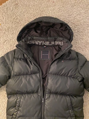 Svart pufferjacka Tommy Hilfiger - Svart pufferjacka från Tommy Hilfiger med huva och broderad logga på insidan av kragen. Jackan har quiltad design och diskret logotyp på bröstet. Perfekt för kalla dagar och har en clean, modern look.  väldigt skön och perfekt för vinterdagarna💫 storlek xs till s