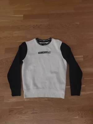 Vit och svart Hollister sweatshirt S - Snygg sweatshirt från Hollister i storlek S. Tröjan är vit med svarta ärmar och har Hollister-logga broderad på bröstet samt tryckt text runt halsringningen. Mjuk insida och klassisk passform, perfekt för chill dagar. Materialet är bomull och polyester.