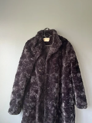 Mörkgrå pälsjacka från Mywear Woman - Säljer en fluffig och lång pälsjacka från Mywear Woman Outerwear Collection i storlek L. Jackan är mörkgrå, har bred krage och stängs med knappar framtill. Perfekt för dig som vill ha en mysig och snygg look under kalla dagar.