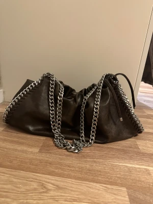 Brun axelväska med kjedjerem - Snygg brun axelväska från Gina Tricot i läderimitation med grova silverfärgade kedjor längs kanterna och som axelrem. Väskan har snörning upptill där man kan justera storleken och den har även ett innerfack med dragkedja. Den är oanvänd då jag har använt andra väskor! Köpt på asos för 700kr. 