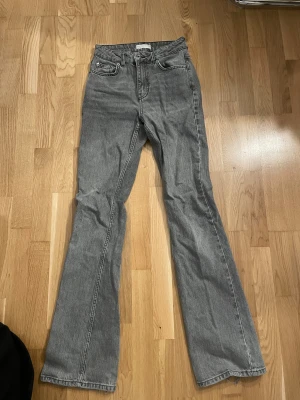 Grå bootcut jeans med mellan hög midja - Snygga grå jeans med bootcut passform och hög midja. Klassisk femficksdesign och dragkedja med knapp framtill. Jeansen har en rak och lite utsvängd siluett som ger en cool vibe. Perfekta för dig som gillar retrostil och vill sticka ut.