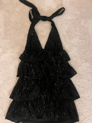 Svart glittrig volangklänning med knyt - Supersnygg svart kortklänning med glittriga paljetter och flera lager volanger. Djup v-ringning och knyt i nacken som ger en öppen rygg. Har bara använt 1 gång! De är bara och skriva om du har några funderingar!💗