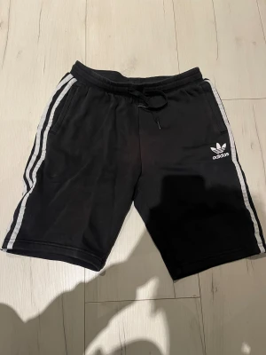 Svarta Adidas shorts med vita ränder - Svarta shorts från Adidas med klassiska vita ränder längs sidorna och Adidas-logga framtill. Mjuk resår i midjan med snörning för skön passform. Perfekta för träning eller chill dagar. Tillverkade i bomullsmix för extra komfort.