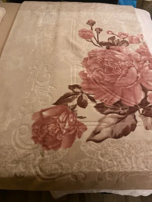 Beige blommig blanket - Säljer en beige blanket med stort blommigt mönster i rosa och bruna toner. Den har en mjuk och fluffig känsla, perfekt för att hålla värmen. 