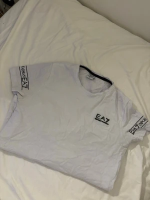 Vit EA7 t-shirt från Armani - Snygg vit t-shirt från Armani EA7 med svart logga på bröstet och tryck på båda ärmarna. Klassisk rund hals och korta ärmar. Perfekt för dig som gillar stilrena och trendiga märkesplagg. Materialet är mjuk bomull som känns skönt mot huden.