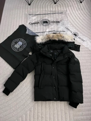 Svart pufferjacka från Canada Goose - Säljer en svart pufferjacka från Canada Goose med avtagbar huva och snygg fuskpälsdetalj. Jackan har flera fickor, ribbade muddar och klassisk logga på ärmen. Perfekt för kalla dagar och riktigt stilren look.