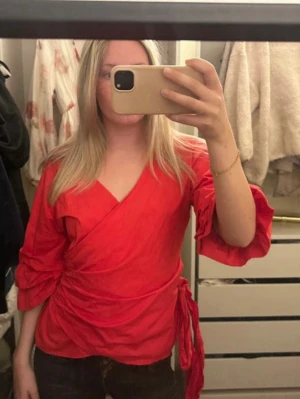 Röd omlottblus med knytband - Säljer en snygg röd omlottblus med v-ringning och trekvartsärmar med volangdetaljer. Blusen har knytband i midjan som ger en fin passform och är tillverkad i ett lätt tyg, perfekt för vår och sommar.