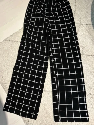 Svarta rutiga wide byxor med resår - Svarta byxor med vitt rutmönster och breda ben. De har en bekväm resår i midjan och är tillverkade i ett mjukt tyg som ger en skön känsla. Passformen är loose och stilen är wide, vilket gör dem riktigt chill och trendiga.