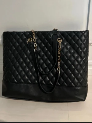 Svart quiltad axelväska med kedja - Stor svart axelväska med quiltat mönster och snygga guldfärgade kedjedetaljer på axelremmarna. Väskan är tillverkad i skinnimitation och har en rymlig insida med fack och dragkedja. Perfekt för dig som vill ha plats för allt och samtidigt se chic ut.