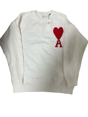 Vit Ami Paris stickad tröja - Säljer en vit stickad tröja från Ami med ett rött hjärta och bokstaven A broderat på bröstet. Tröjan har rund hals, ribbade muddar och är långärmad. Perfekt för dig som gillar stilrena detaljer och vill ha något som sticker ut lite.
