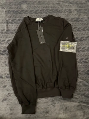Svart sweatshirt Stone Island - Svart långärmad sweatshirt Stone Island med klassisk rund hals och ribbade muddar. Tröjan har den ikoniska Stone Island-patchen med gul brodyr som fästs med knappar på ärmen. Mjuk bomullskvalitet och stilren design, perfekt för dig som gillar streetwear.