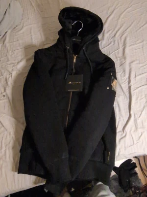 Moose Knuckles hoodie/jacka - Säljer en svart pufferjacka från Moose Knuckles i storlek XS. Jackan har huva med fuskpäls, guldfärgade detaljer och dragkedja framtill. Innerfodret är quiltat och jackan har en ikonisk logga på ärmen. Perfekt för kalla dagar och riktigt snygg streetstil. ÄKTA!