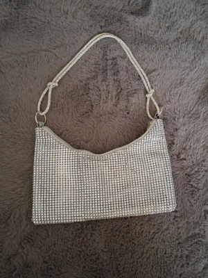 Glittrig silver axelväska - Säljer en snygg axelväska i silver med glittriga stenar över hela väskan. Den har ett rektangulärt format och ett matchande axelband med knutdetaljer. Väskan stängs med dragkedja och är perfekt för att lyfta vilken outfit som helst.