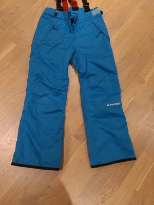 Blå skibyxor från Everest, stl 146 - Säljer ett par blå skibyxor från Everest i storlek 146. Byxorna har svarta och röda hängslen, dragkedjor vid fickorna och är fodrade för extra värme. Perfekta för vinterns alla äventyr i snön!