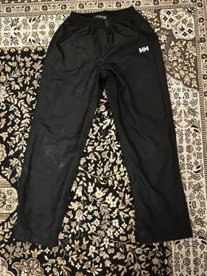 Svarta täckbyxor Helly Hansen - Svarta täckbyxor från Helly Hansen med elastisk midja och HH-logga på framsidan och inuti byxan. Byxorna har raka ben och justerbara benslut med kardborre. Byxorna är fodrade och lämpar sig för kallt väder.