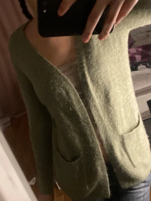 Grön stickad kofta med fickor - Mysig grön stickad kofta med lång ärm och två praktiska fickor framtill. Koftan har en öppen design utan knappar och är perfekt att slänga på sig över en topp. Den har en loose passform och känns riktigt skön mot huden.