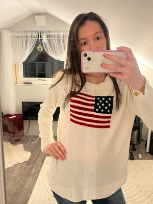 Vit stickad tröja med USA-flagga - Vit stickad tröja med en stor amerikansk flagga på bröstet. Tröjan har rund halsringning och långa ärmar. Perfekt för dig som vill ha en chill och snygg look med lite preppy känsla. Obs det står ingen storlek på men passar som s-m!