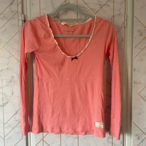 Rosa ribbad långärmad topp Odd Molly - Supersöt rosa ribbad topp från Odd Molly med lång ärm, v-ringning med vit spetskant och liten rosett framtill. Toppen har tryckt logga på ena ärmen och är i mjuk bomull