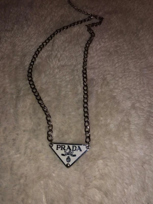 Prada silverfärgad halsband - Snyggt halsband från Prada med en kraftig silverfärgad kedja och en ikonisk triangelformad platta med logotypen framtill. Plattan är vit med svarta detaljer och kedjan har en glansig finish som ger en lyxig vibe. Perfekt accessoar för att lyfta din outfit.