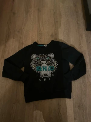 Svart Kenzo sweatshirt med tiger - Säljer en svart sweatshirt från Kenzo med ikonisk broderad tiger i vitt, grått och turkost på bröstet. Det står att storleken är s men den är xs så du vet. Har ett litet hål vid sidan av magen men är perfekt statement-plagg för dig som gillar streetstyle och vill sticka ut.
