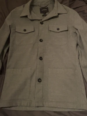 Grå overshirt från Dressmann S - Stilren grå overshirt från Dressmann i slim fit-modell. Jackan har klassisk krage, fyra stora fickor framtill och svarta knappar hela vägen. Perfekt att slänga på över en hoodie eller t-shirt för en chill look.