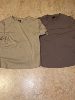 Två basic t-shirts från SHEIN XS - Säljer två stilrena t-shirts från SHEIN i storlek XS. Ena är beige och den andra är brun, båda med rund hals och korta ärmar. Materialet är mjukt och känns som polyester, perfekt för en chill och enkel look.