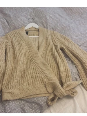 Beige omlott kofta från Y.A.S - Säljer en beige stickad kofta från Y.A.S med omlott-design och knytband i midjan. Koftan har långärmade ärmar och grovstickad struktur som ger en mysig känsla. Perfekt att slänga på sig när det är lite kyligt ute.