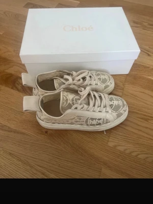 Beige spets-sneakers från Chloé - Säljer ett par snygga beige sneakers från Chloé med spetsdetaljer och tydlig logga på sidan och hälen. Skorna har snörning och platt sula, samt en stor tygflik bak med Chloé-tryck. Perfekta för dig som gillar unika och trendiga detaljer.