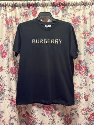 Svart Burberry t-shirt med broderi - Svart t-shirt från Burberry i mjuk bomull, med klassisk rund hals och korta ärmar. Framsidan har ett broderat Burberry London England-tryck i beige och röd ton. Perfekt för dig som gillar stilrena designer med lyxig känsla.