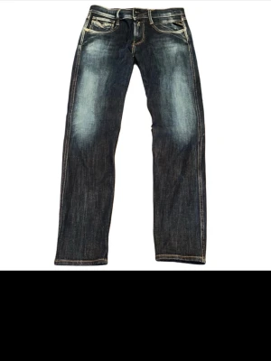 Replay Anbass jeans blå  - Snygga Replay Anbass jeans i mörkblå tvätt med slitningar och kontrastsömmar. Klassisk femficksmodell med raka ben och normal passform. Jeansen har detaljerade bakfickor med broderi och Replay-logga på linningen. Perfekt för dig som gillar stilrena och coola jeans.