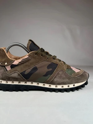 Valentino Garavani Rockrunner sneakers - Säljer ett par Valentino Garavani Rockrunner sneakers i camomönster med olivgröna och bruna toner. Skorna har detaljer i mocka, nitar runt sulan och gröna snören. Snygg chunky sula och coola militärvibbar. Perfekta för dig som vill sticka ut med exklusiva detaljer.