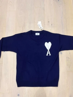 Marinblå stickad tröja från Ami - Snygg marinblå stickad tröja från Ami med ett stort vitt hjärta och bokstaven A på bröstet. Tröjan har rund hals, ribbade muddar och lång ärm. Perfekt för dig som gillar stilrena detaljer och vill ha något som sticker ut lite extra.