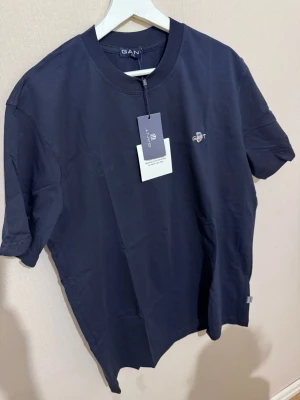 Mörkblå t-shirt från GANT - Snygg mörkblå t-shirt från GANT med diskret logobrodyr på bröstet. Klassisk rund hals och korta ärmar. Tillverkad i mjuk bomull som känns skön mot huden. Perfekt för dig som gillar stilrena och enkla plagg med en touch av premiumkänsla.Den kan passa storlek XL också. Hör av dig vid frågor 