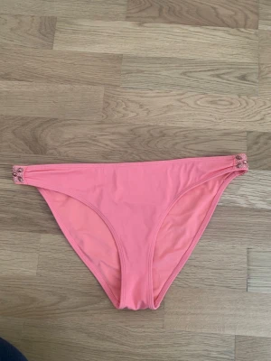 Rosa bikiniunderdel med pärlor från H&M - Säljer en snygg rosa bikiniunderdel från H&M i storlek 40. Modellen har lågt skuren midja och är dekorerad med glittriga pärlor och stenar på sidorna för extra bling. Materialet är mjukt och stretchigt syntet, perfekt för strand eller pool.