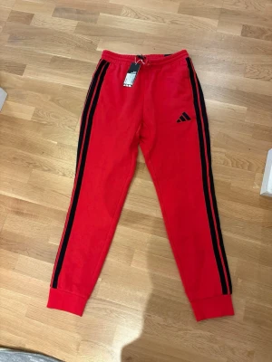 Röda Adidas mjukisbyxor med svarta ränder - Säljer ett par röda mjukisbyxor från Adidas i storlek S. Byxorna har svarta ränder längs sidorna, elastisk midja med snörning och Adidas-logga på benet. Tillverkade i mjukt bomullsmaterial, perfekta för chill eller träning. Oanvända med prislapp kvar.