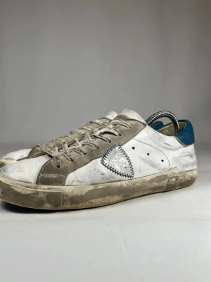 Philippe Model  - Snygga vita sneakers från Philippe Model med grå mockadetaljer och blå häl. Skorna har snörning och en unik broderad patch på sidan. Yttermaterialet är skinn och mocka, med platt sula i gummi och rund tå. Perfekt för dig som gillar grisch och exklusiva märken.