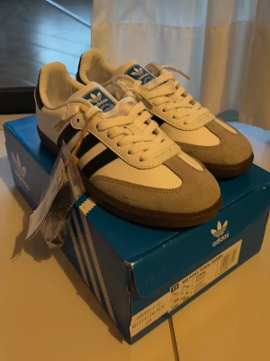 Adidas Samba skor - Adidas Samba sneakers i klassisk design med vit ovandel i skinn, svarta ränder på sidorna och grå mockadetaljer vid tån. Platt brun sula och snörning framtill. Perfekt för dig som gillar retrostil och sportig vibe.