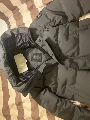 Mörkgrå pufferjacka från Canada Goose - Säljer en mörkgrå pufferjacka från Canada Goose med ikonisk rund patch på ärmen. Jackan har huva, dragkedja och knappar framtill, samt flera praktiska fickor. Materialet är polyester och dun, perfekt för kalla dagar. Stilsäker och riktigt varm.