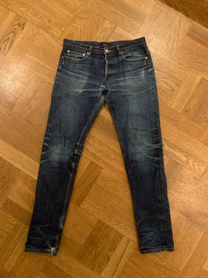 A.P.C. Petit New Standard jeans  - Säljer ett par A.P.C. Petit New Standard jeans i mörkblå tvätt med snygga slitningar. Modellen har smal passform, klassisk femficksdesign och knäppning med silverfärgade knappar. Tillverkade i 100% bomull för en autentisk jeanskänsla. Raw selvedge, använd ett par månader med ett par tvättningar