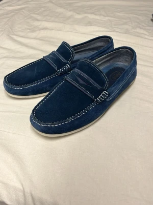 Marinblå mockaloafers med vit sula - Snygga marinblå loafers i mocka med kontrasterande vita sömmar och platt vit sula. Skorna har klassisk rund tå och stilren design som passar till jeans eller chinos. Perfekt för dig som gillar en chill och stilren look.