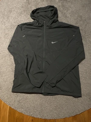 Svart vindjacka från Nike med huva - Snygg svart vindjacka från Nike med huva och hel dragkedja framtill. Jackan har en stilren design med Nike-logga på bröstet och praktiska fickor på sidorna. Perfekt för blåsiga dagar och sportiga outfits.